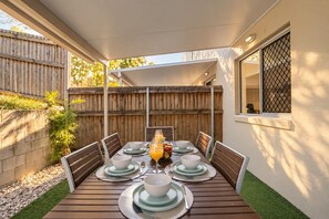 Une terrasse couverte avec une table en bois, mise pour quatre personnes, entourée de chaises, et une fenêtre sur la droite.
