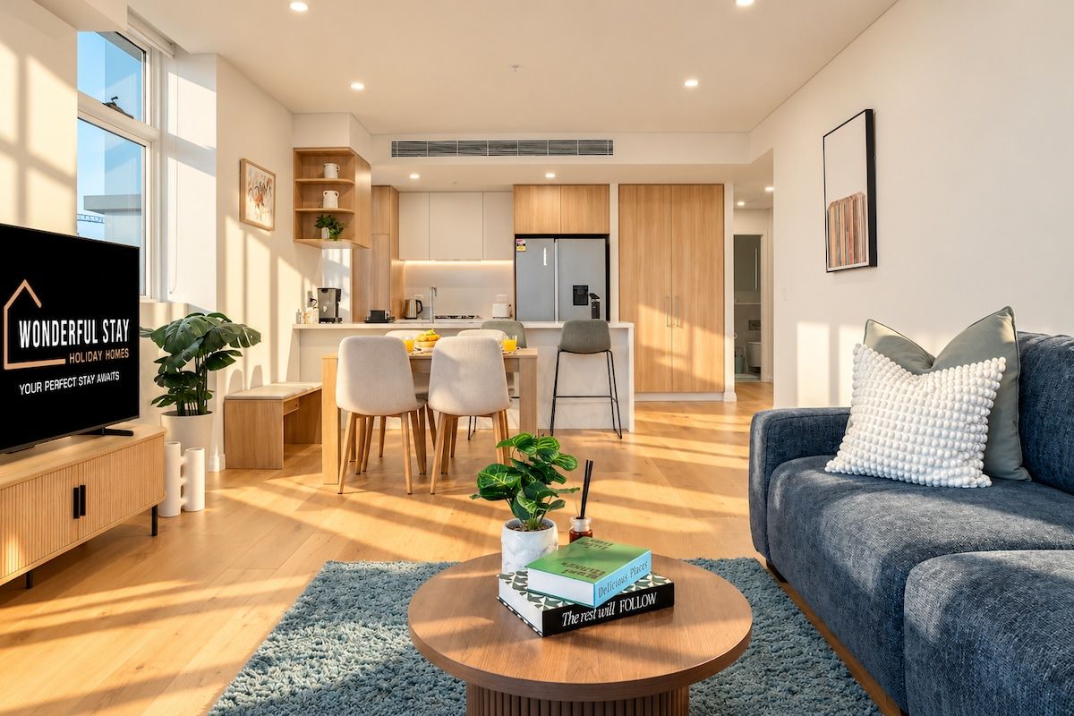 A modern living room with a flat-screen TV, a blue sofa, a coffee table with books, and a dining area with chairs and a kitchen in the background.