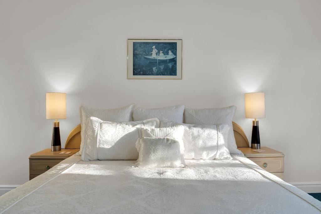 A neatly made bed with white linens and pillows, flanked by two bedside tables with lamps, in a room with a simple white wall and a framed picture above the bed.