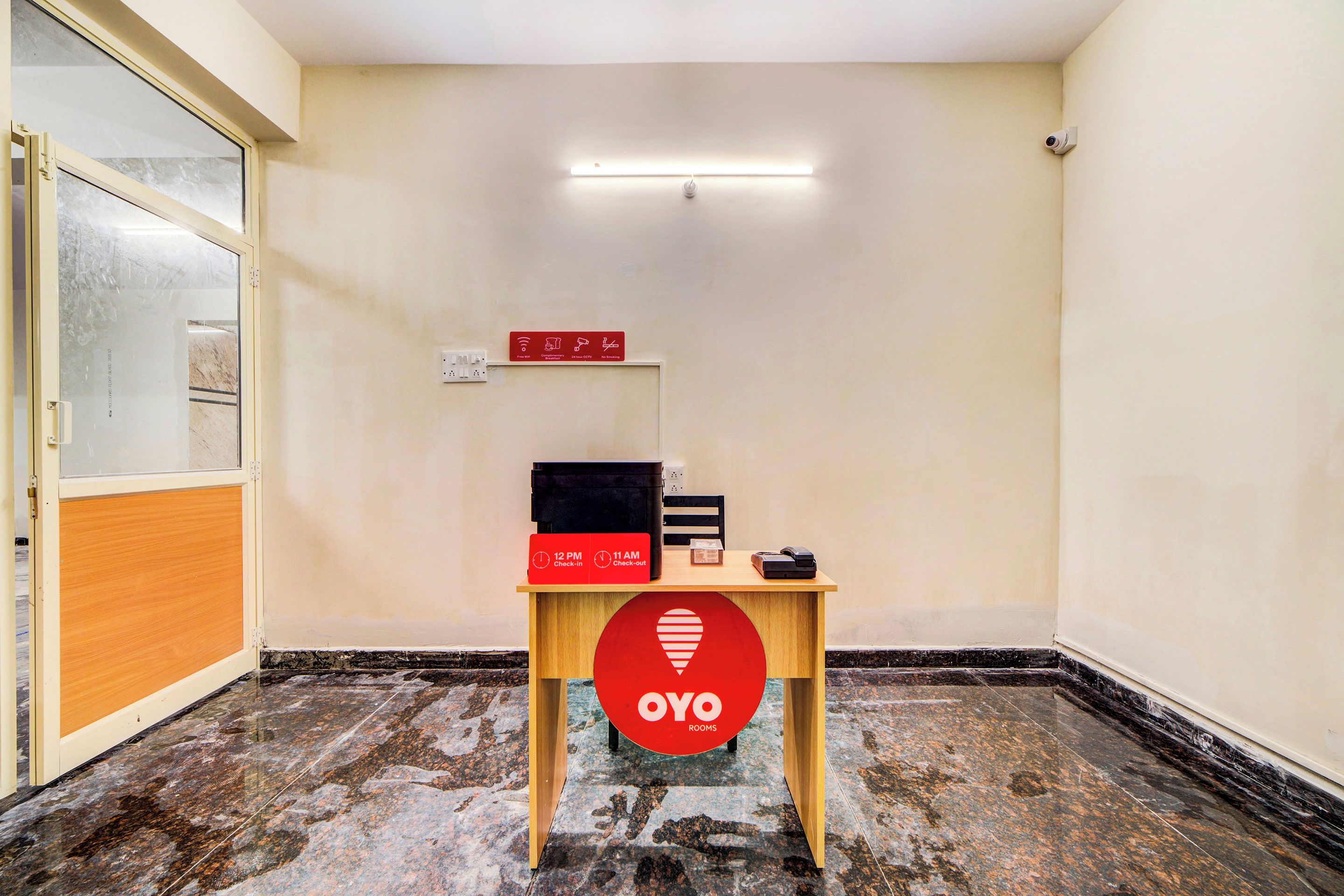 A hotel room with a check-in/out desk, a security camera, and a sign indicating check-in at 12 PM and check-out at 11 AM.