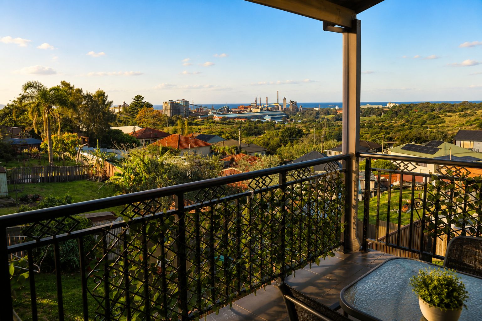 A balcony with a metal railing overlooking a residential area with houses, greenery, and a distant view of industrial structures.