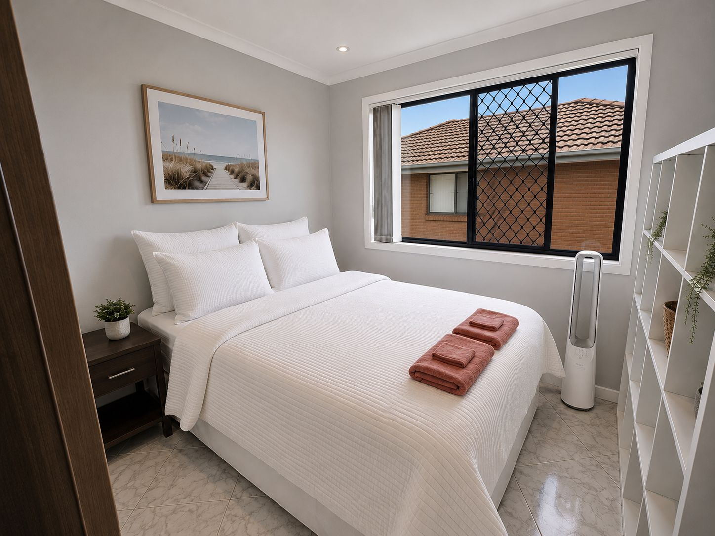 A bedroom with a bed, a nightstand, a potted plant, a window with a view of a building, and a wall-mounted picture.