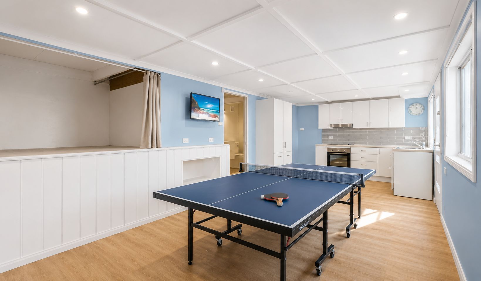 A room with a ping pong table, a TV, and a kitchen area.