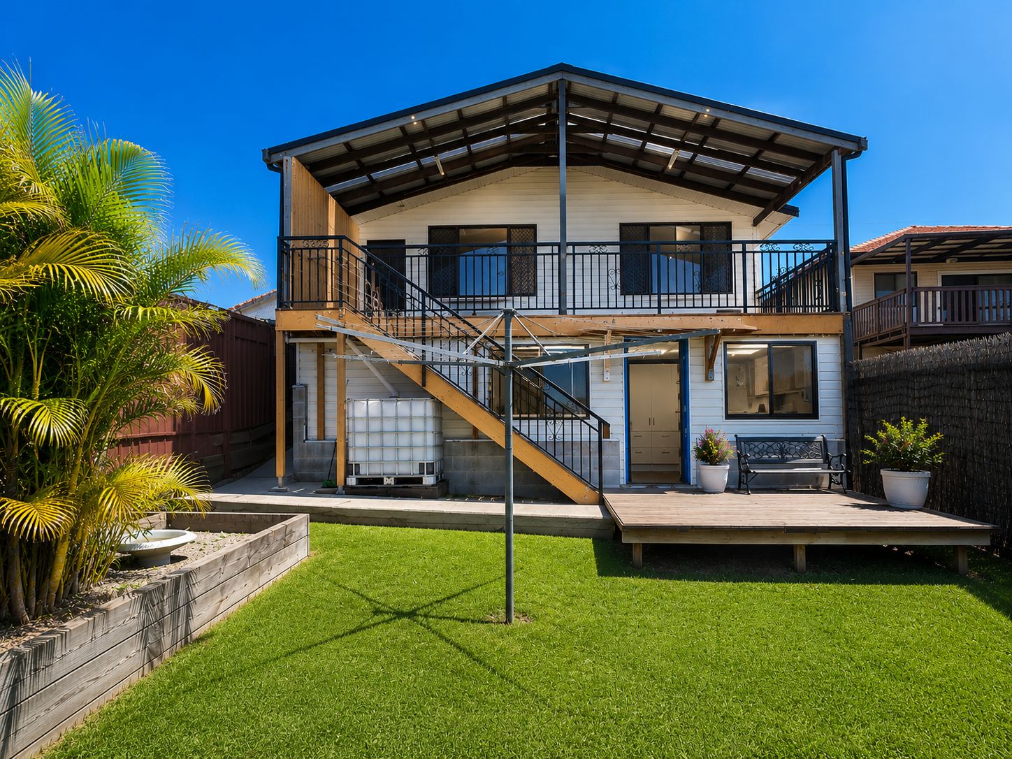 A two-story house with a covered porch, a staircase, and a fenced yard with grass and palm trees.