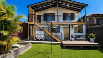 A two-story house with a covered porch, a staircase, and a fenced yard with grass and palm trees.