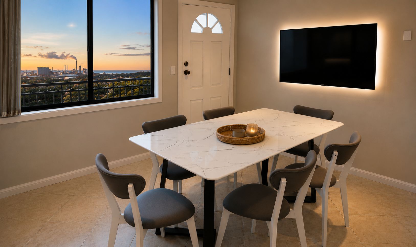 A dining area with a white marble table, six chairs, a flat-screen TV mounted on the wall, and a view of an industrial area through a window.