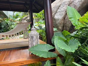 Um deck de madeira com uma rede, um candeeiro e plantas verdes exuberantes.