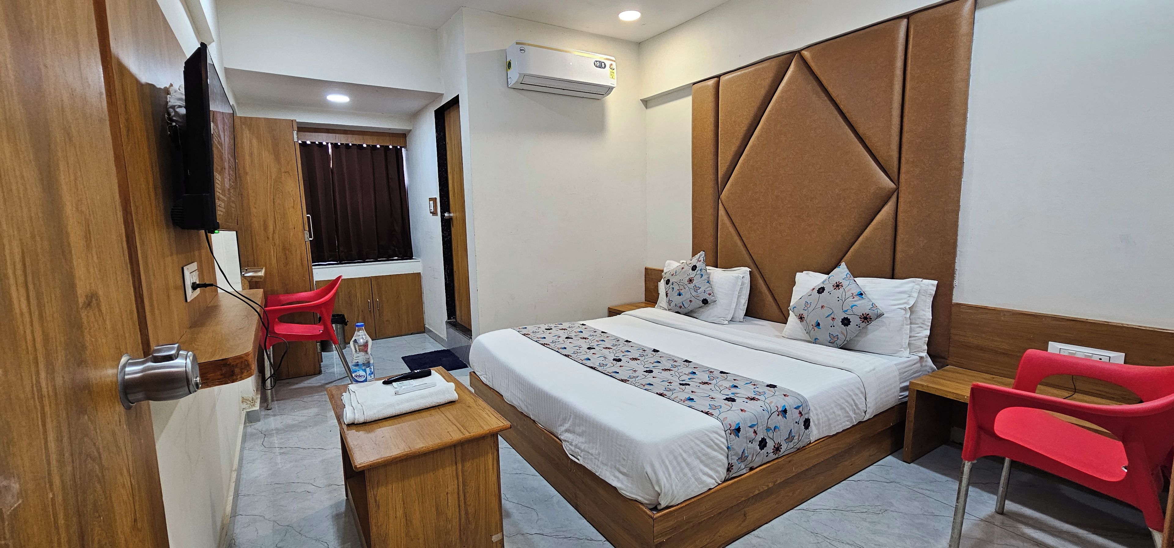 A hotel room with a bed, a desk, a chair, a TV, and an air conditioning unit.
