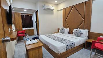 A hotel room with a bed, a desk, a chair, a TV, and an air conditioning unit.