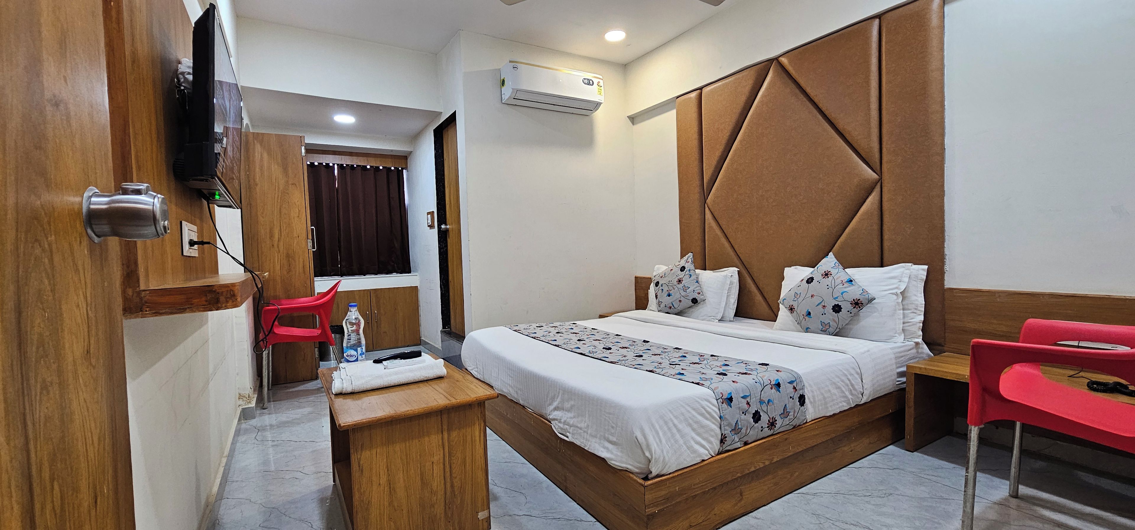 A hotel room with a bed, bedside table, red chairs, a wooden wardrobe, and an air conditioning unit.