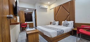 A hotel room with a bed, bedside table, red chairs, a wooden wardrobe, and an air conditioning unit.