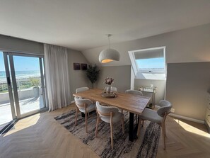 Een eetruimte met een houten tafel, zes stoelen, een hanglamp en uitzicht door grote ramen.