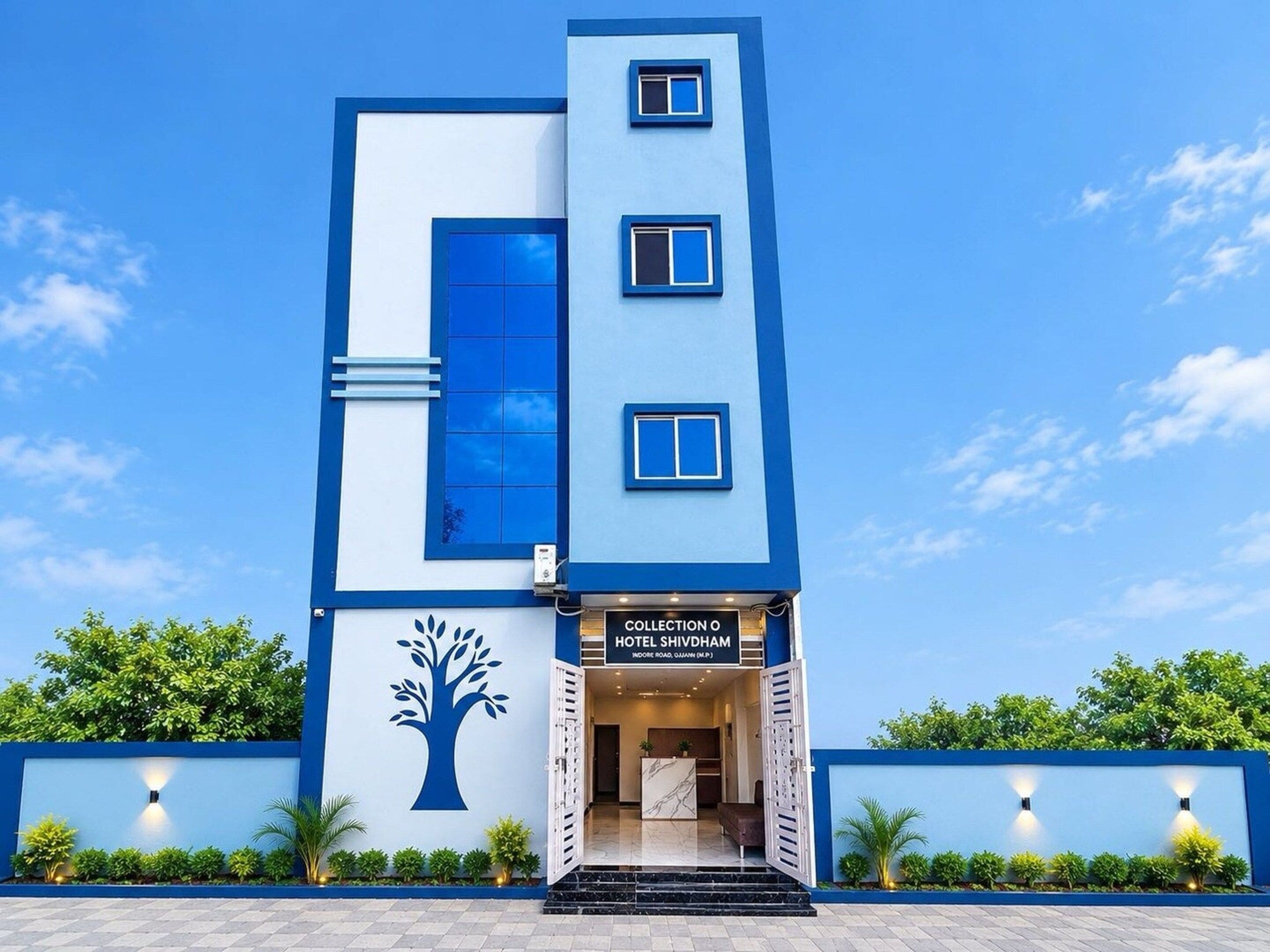Un hotel moderno con fachada azul y blanca, un logotipo de árbol y un cartel que dice 'Collection O Hotel Shivdham'.