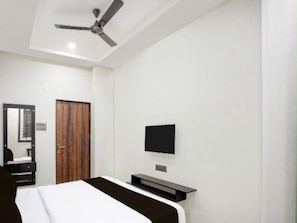Ein Hotelzimmer mit einem Bett, einem an der Wand befestigten Fernseher, einem Deckenventilator und einer Holztür.
