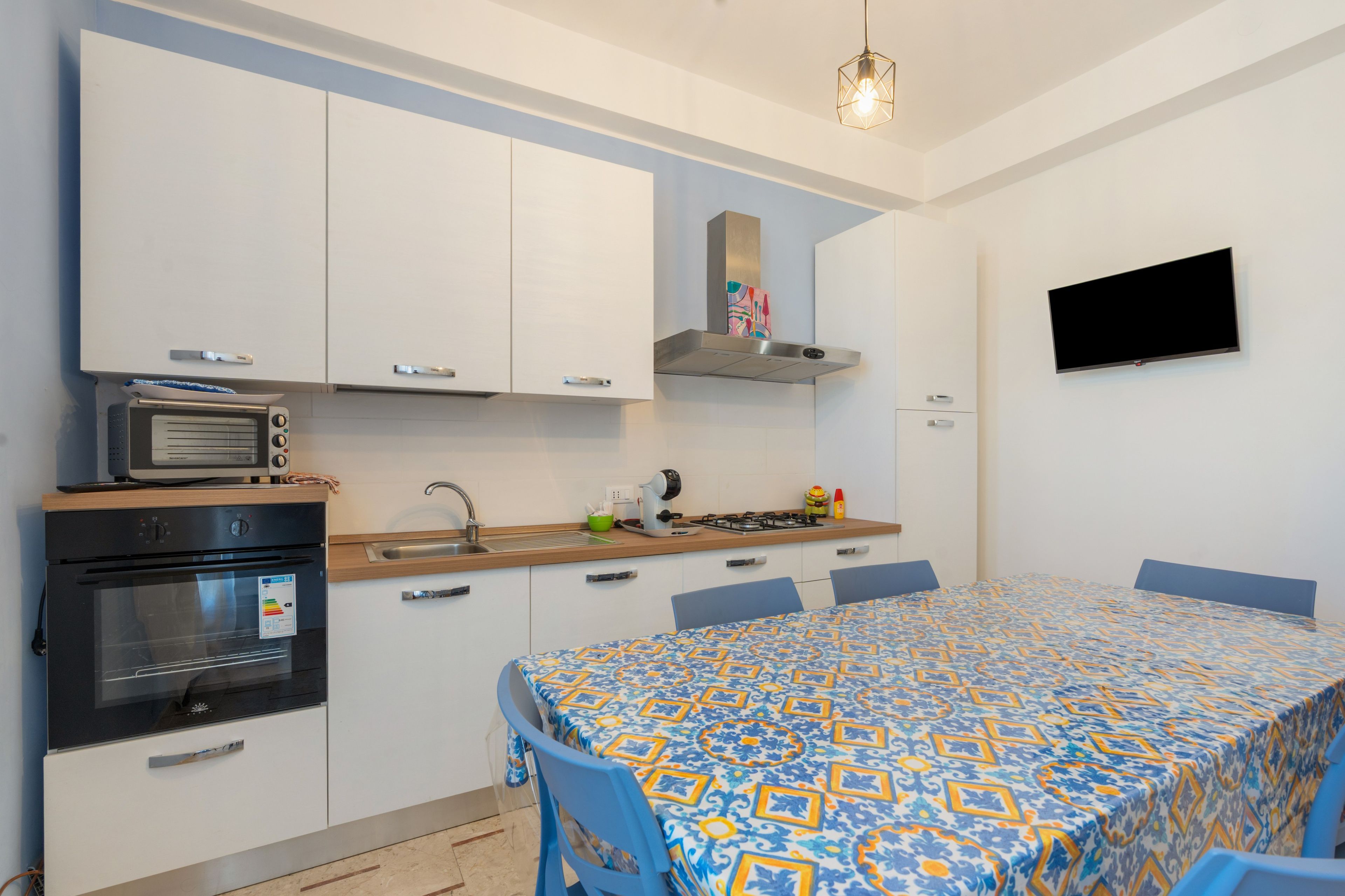 Una cocina compacta con electrodomésticos modernos, un comedor con mantel estampado y un televisor de pantalla plana montado en la pared.