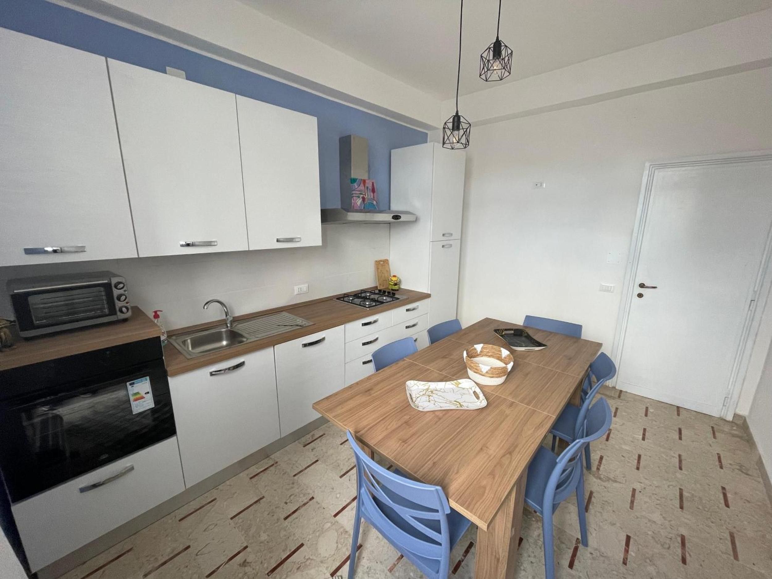 Une cuisine compacte avec des meubles blancs, un plan de travail en bois, un four encastré et un coin repas avec des chaises bleues.