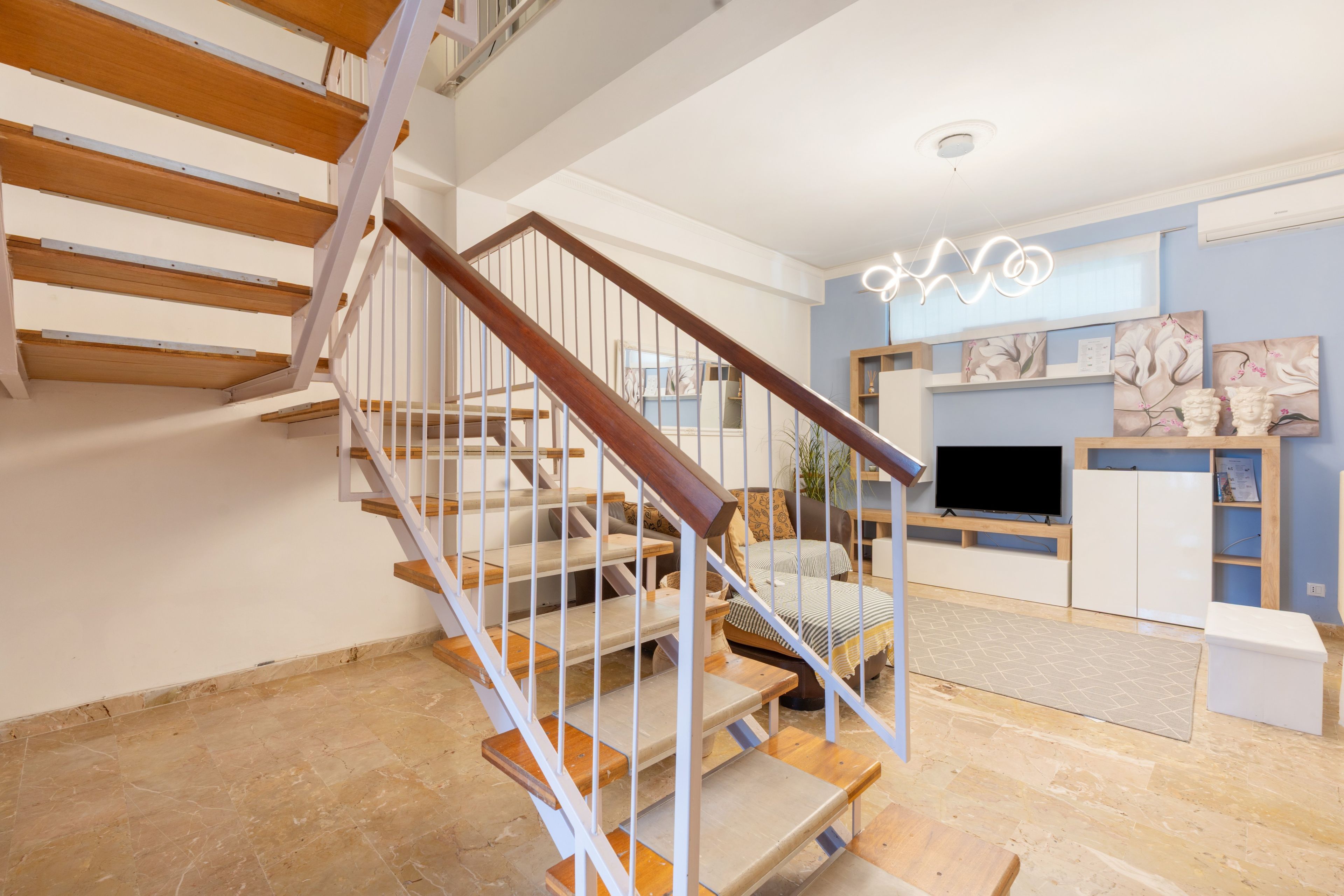 A staircase with wooden steps and white railings, leading to a living area with a flat-screen TV, a sofa, and decorative wall art.