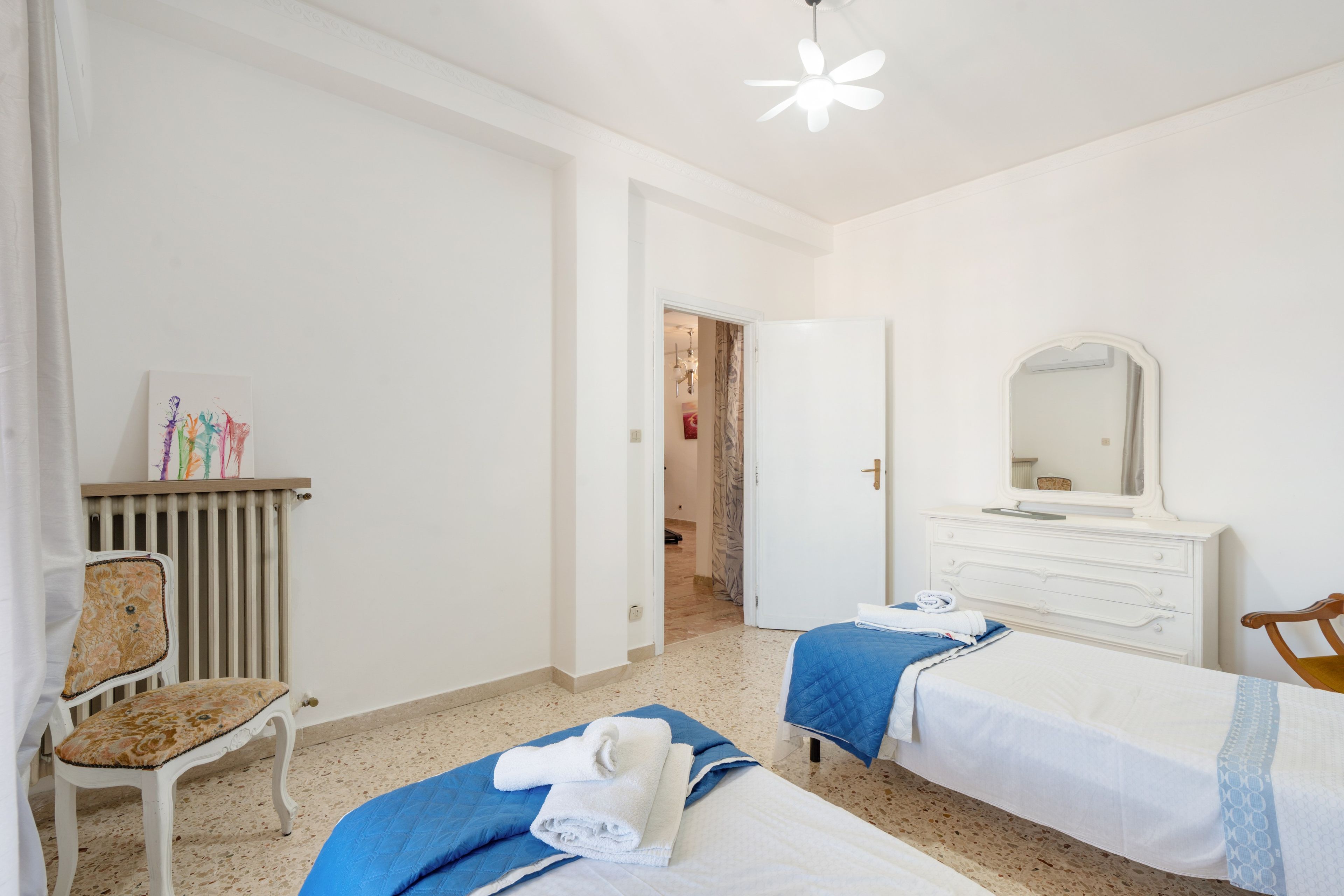 Een hotelkamer met twee bedden, een stoel, een radiator, een spiegel en een deur naar een andere kamer.