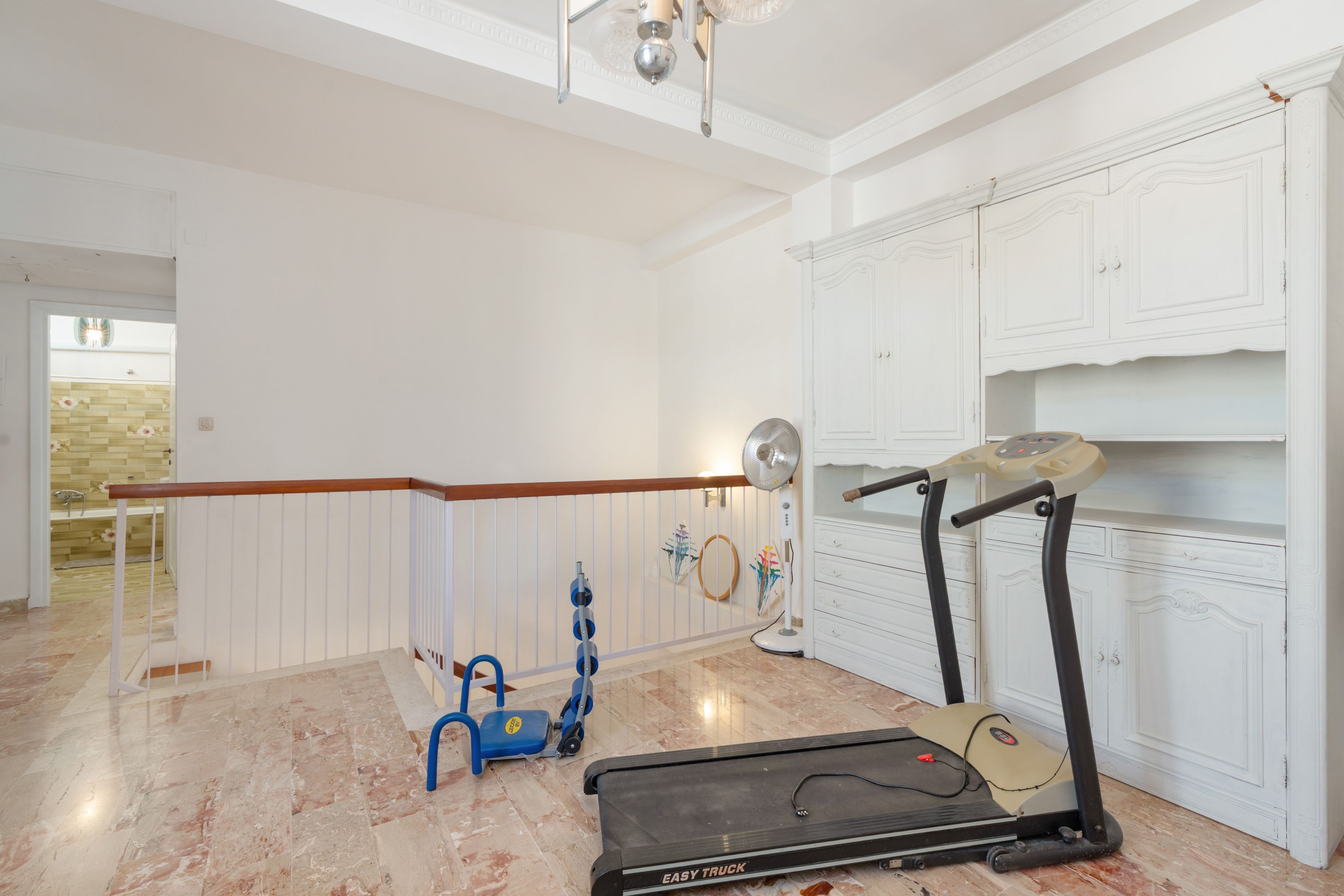 Una habitación amplia con techo blanco, baranda de madera, caminadora, ventilador y pared decorativa.