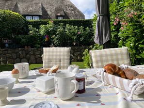 An outdoor dining area with a table set for breakfast, including bread, eggs, and jam, with a thatched-roof building in the background. - Deichhof Apt. 23 (Dunsum)