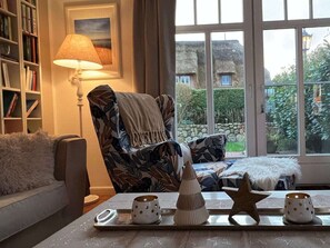A cozy living room with a patterned armchair, a side table with decorative items, a bookshelf, and a window with a view of greenery and buildings. - Deichhof Apt. 23 (Dunsum)