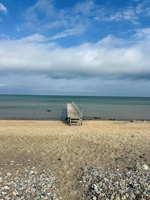 A beach with a wooden walkway leading to the water, pebbles in the foreground, and a calm sea under a cloudy sky. - Holiday home with enclosed garden and terrace (Græsted)