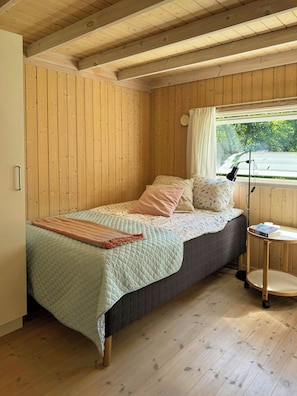 A single bed with a quilt and pillows, a bedside table with a lamp, and a window with curtains. - Holiday home with enclosed garden and terrace (Græsted)