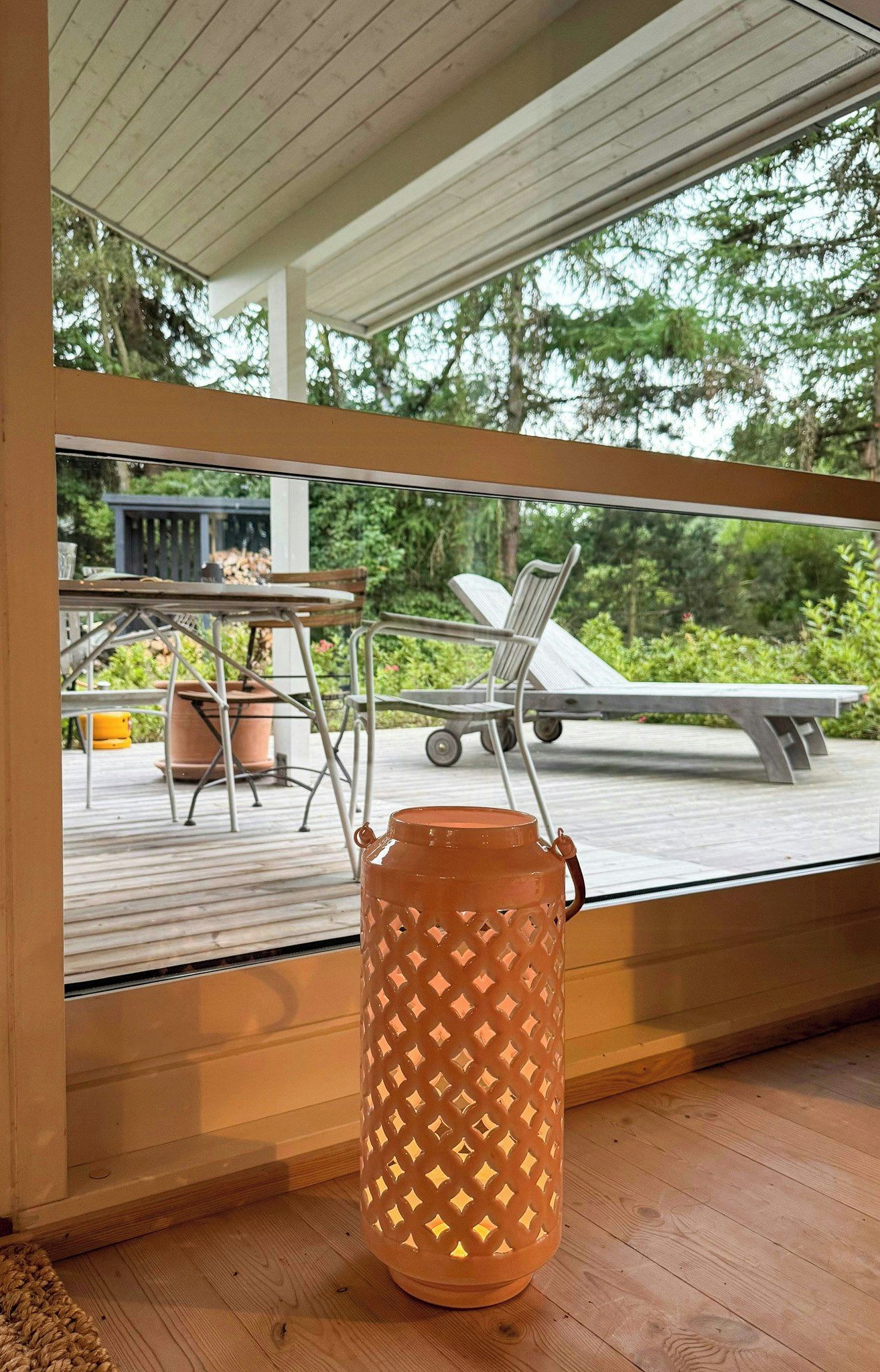 A modern outdoor seating area with a table and chairs, a large planter, and a view of a deck and trees.