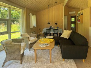 A cozy living room with wicker furniture, a dark sofa, and a dining table with a patterned tablecloth. - Holiday home with enclosed garden and terrace (Græsted)