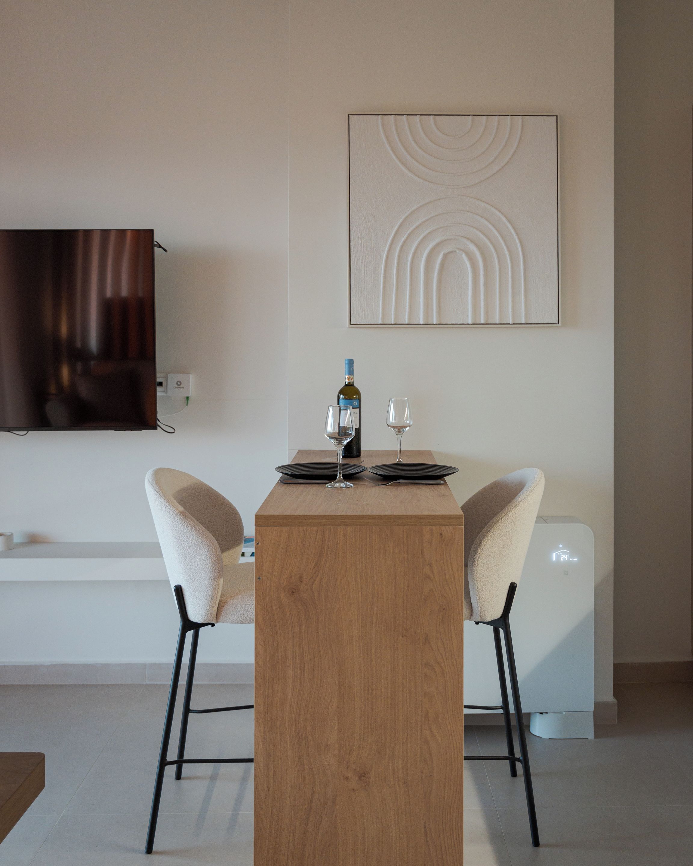 A modern dining area with a wooden table, two chairs, a wine bottle, and a glass. A flat-screen TV is mounted on the wall above the table.