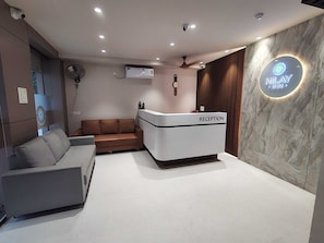 A reception area with a reception desk labeled 'RECEPTION,' a brown sofa, a grey sofa, a wall-mounted clock, and a sign that reads 'NILAY INN.' - Classic Nilay Inn (Pandharpur)