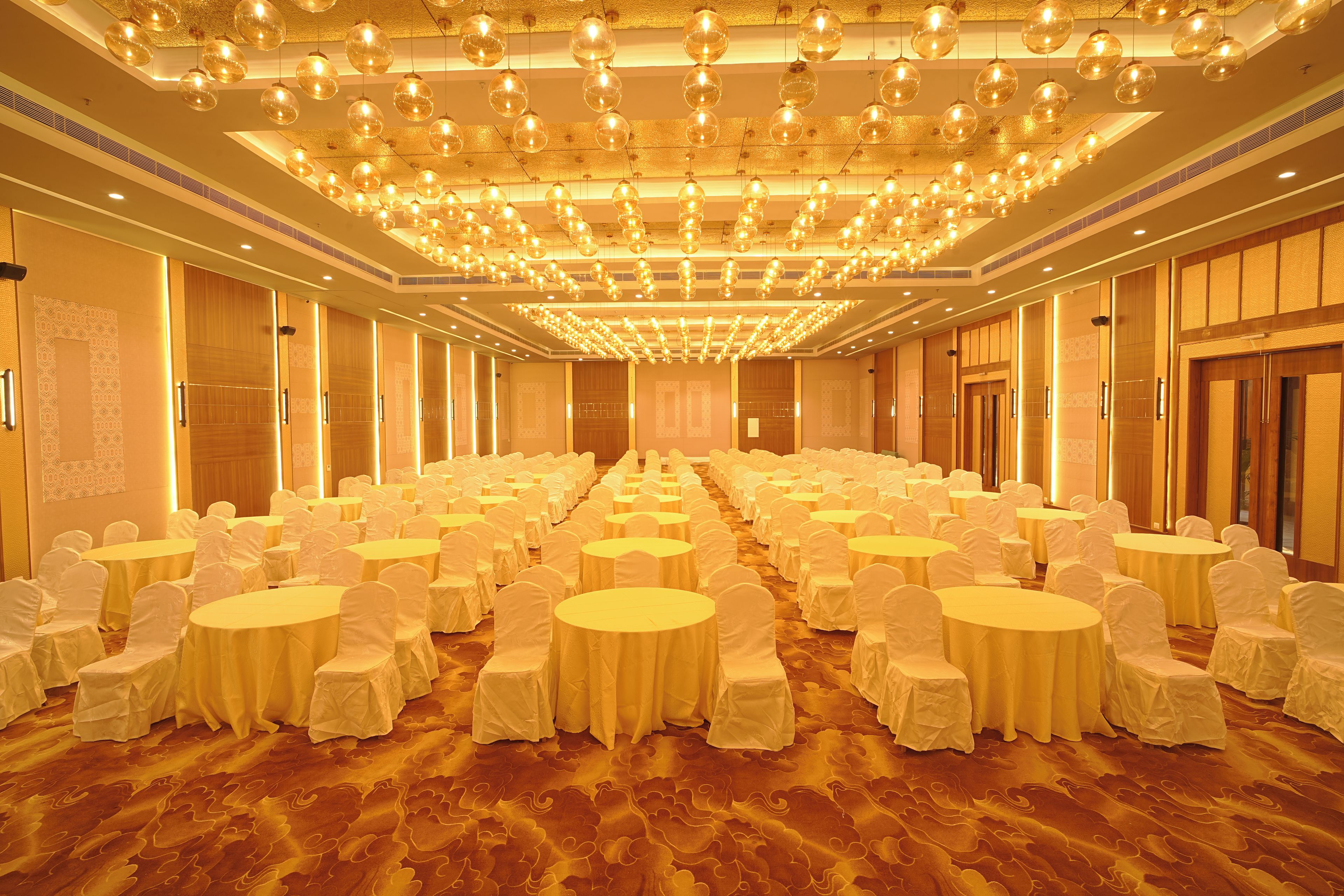 A banquet hall with round tables and chairs, a patterned carpet, and a ceiling with hanging spherical lights.