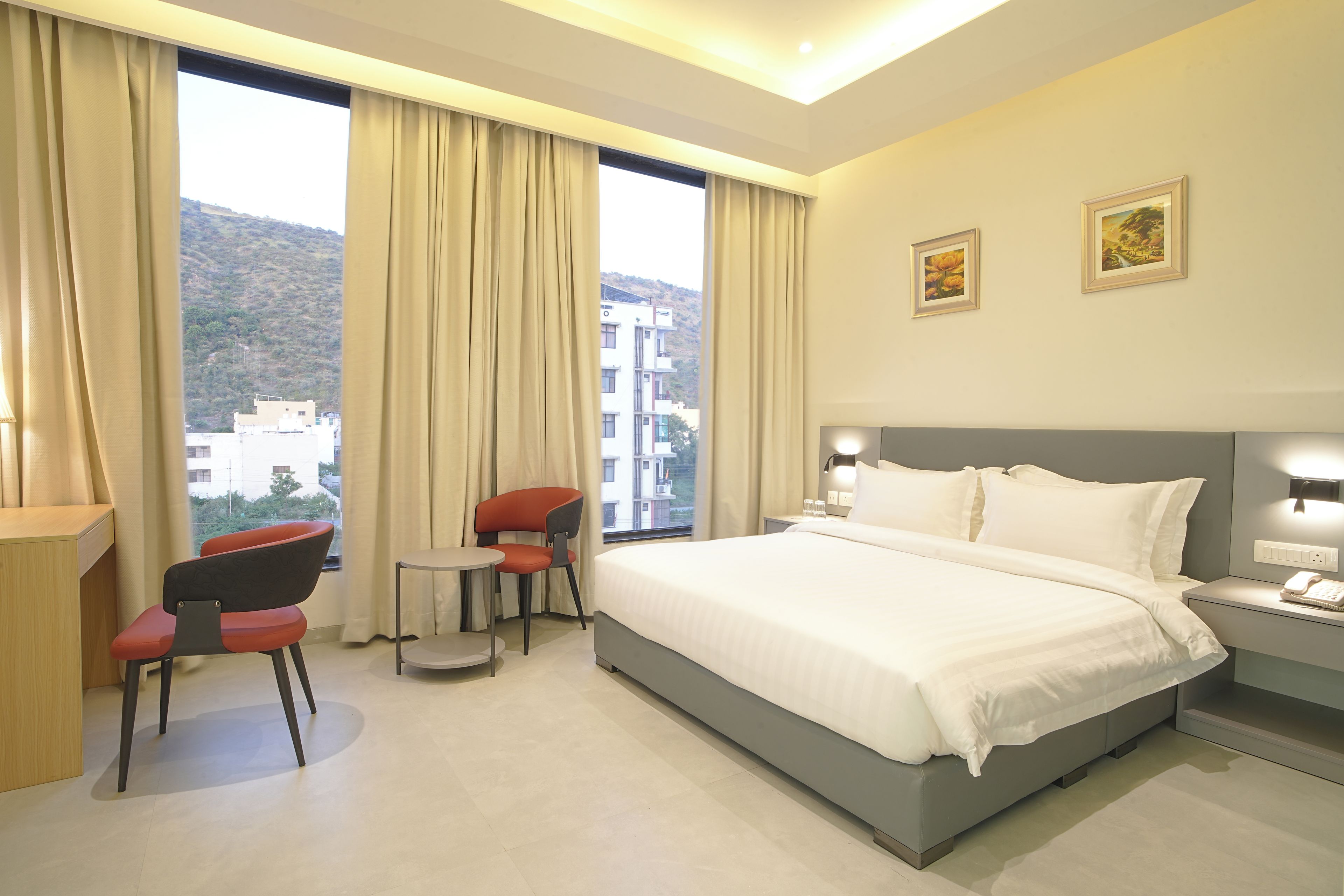 A hotel room with a bed, two chairs, a small table, a nightstand with a phone, and two paintings on the wall.