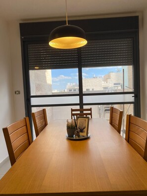 A dining area with a wooden table, six chairs, a pendant light, and a view of buildings through a large window with blinds. - Mesilat Yesharim Penthouse  (Jerusalem)