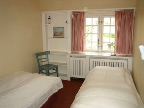 A room with two single beds, a chair, a radiator, a window with curtains, and a small shelf with a picture. - Waldow im Ulmenhof 5 (Nieblum)