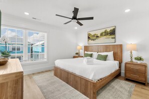 A bedroom with a wooden bed, a ceiling fan, a window with blinds, a bedside table with a lamp, and a painting on the wall. - The Blue Seeker Steps to Beach Pet Friendly (Myrtle Beach)