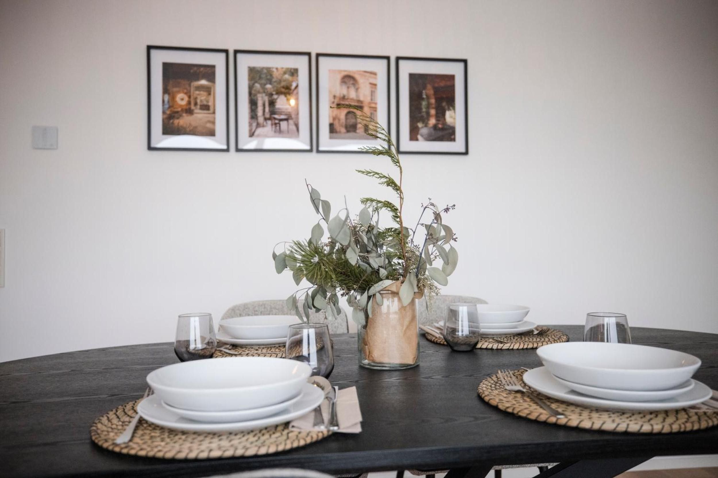A dining table set with white plates, glasses, and a vase with green foliage.