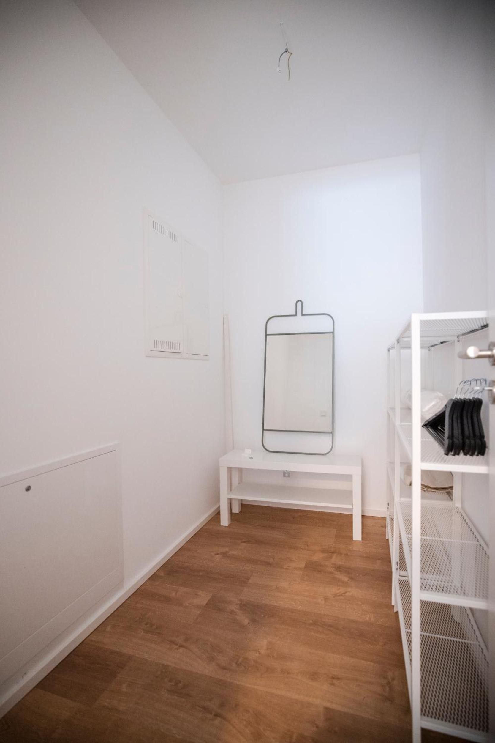 A minimalist room with a wooden floor, a white bench, a mirror, and a white shelving unit.