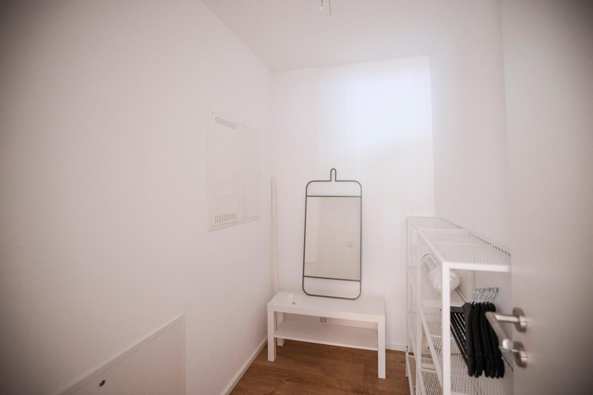 A small, white room with a wooden floor, a white bench, a mirror on the wall, and a white shelving unit with clothes.