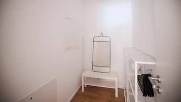A small, white room with a wooden floor, a white bench, a mirror on the wall, and a white shelving unit with clothes.