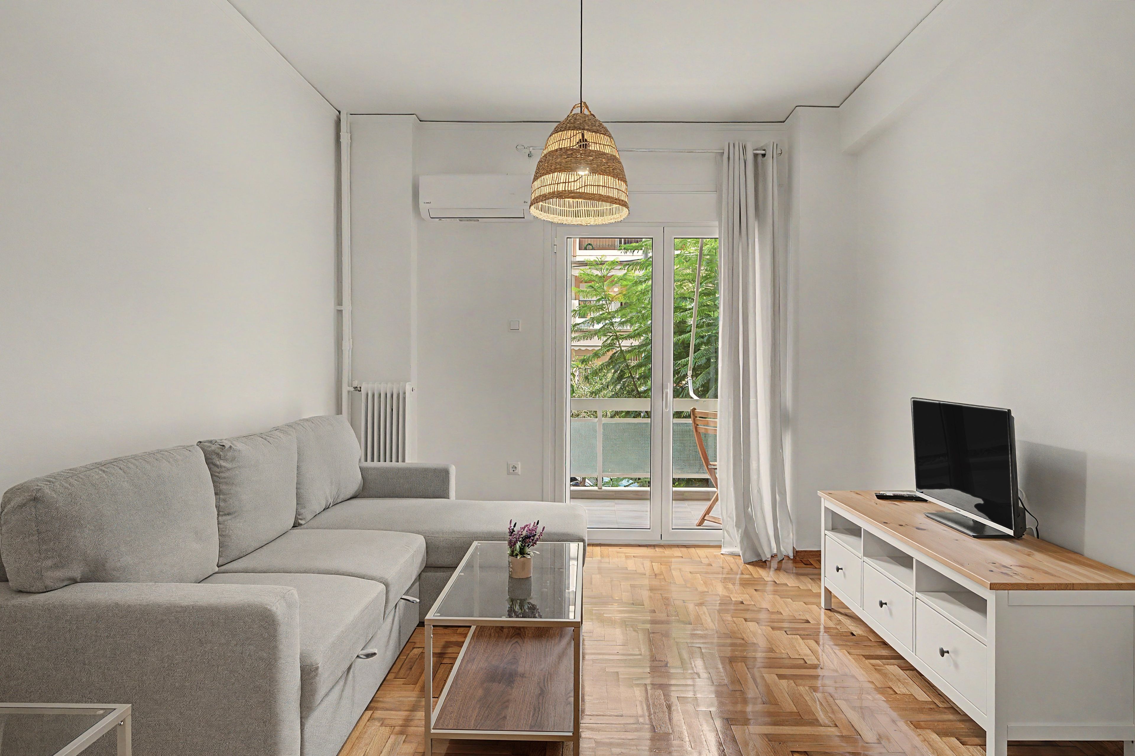 A modern living room with a grey sofa, a glass coffee table, a wooden TV stand, and a large window with a view of greenery.