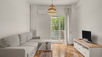 A modern living room with a grey sofa, a glass coffee table, a wooden TV stand, and a large window with a view of greenery.