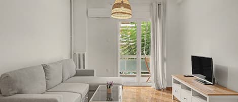 Una sala de estar moderna con un sofá gris, una mesa de centro de vidrio, un mueble de televisión de madera y un ventanal con vistas a zonas verdes.