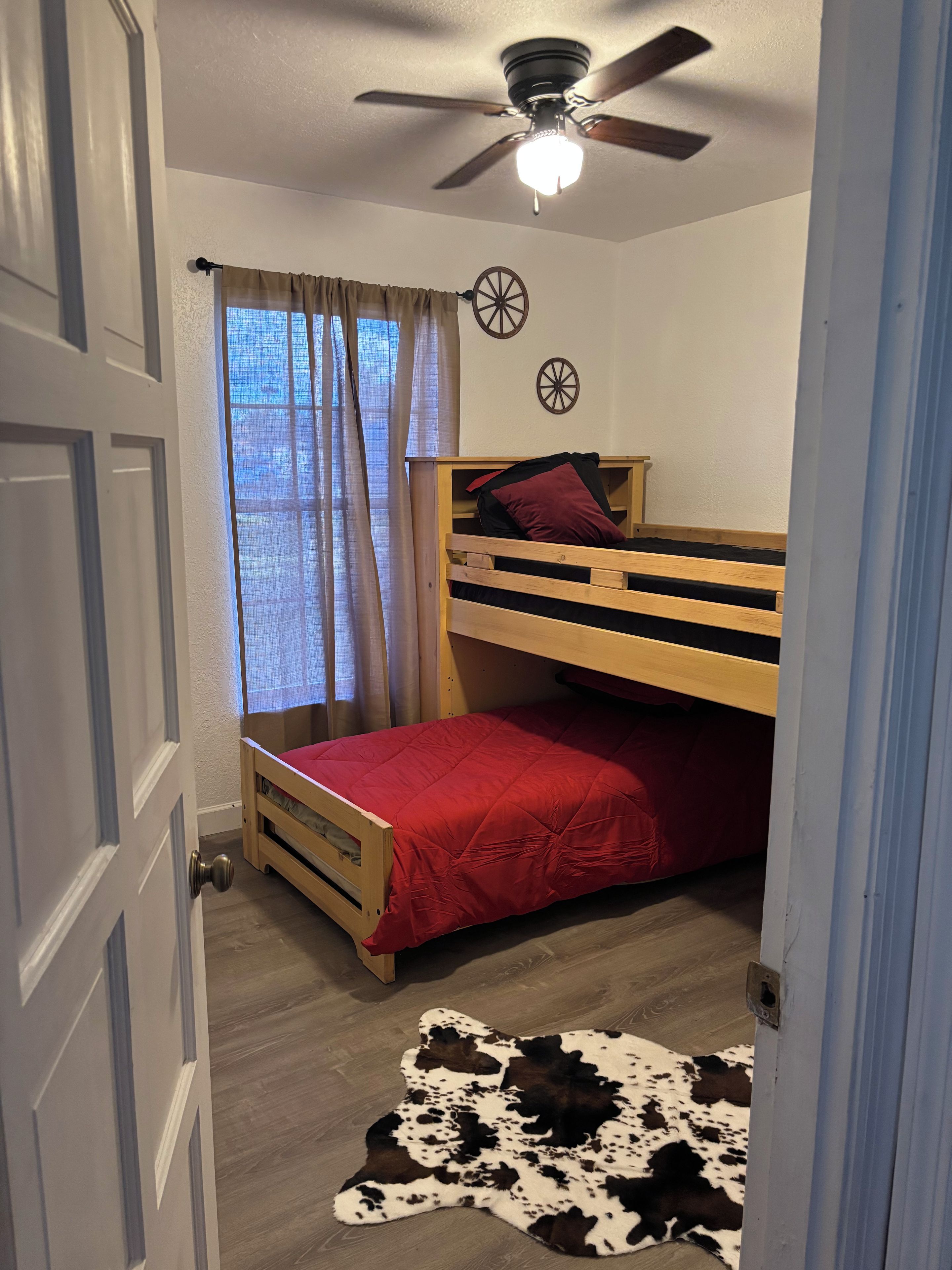 Una habitación con literas, ventilador de techo, alfombra de cuero y una ventana con cortinas.