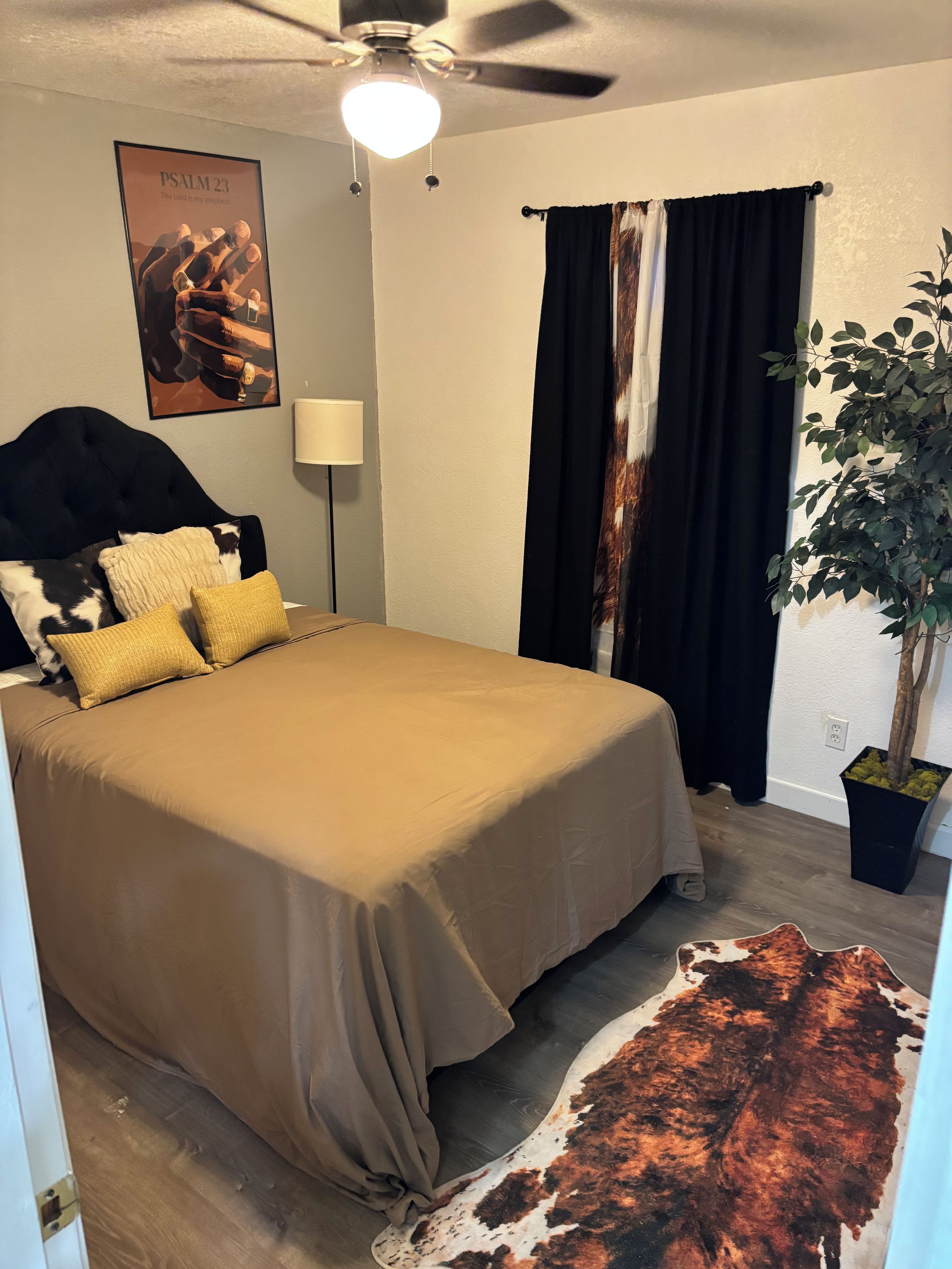 Ein Schlafzimmer mit einem Bett, einem Deckenventilator, einer Wandlampe, einem Poster, einer Pflanze und einem Kuhfellteppich.