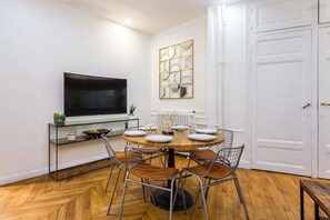 A dining area with a round table set for four, surrounded by wire mesh chairs. A flat-screen TV is mounted on the wall above the table. - Welcoming Apartment -1br/6p - Auteuil / Passy (Paris)