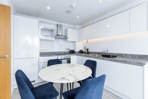 Une cuisine moderne avec des meubles blancs, un four encastré, un four à micro-ondes et une cuisinière à gaz. On y trouve une table ronde en marbre, entourée de chaises bleues.