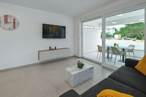 Una sala de estar moderna con un sofá, una mesa de centro, un televisor y un ventanal corredera que da a un espacio exterior con una mesa y sillas.