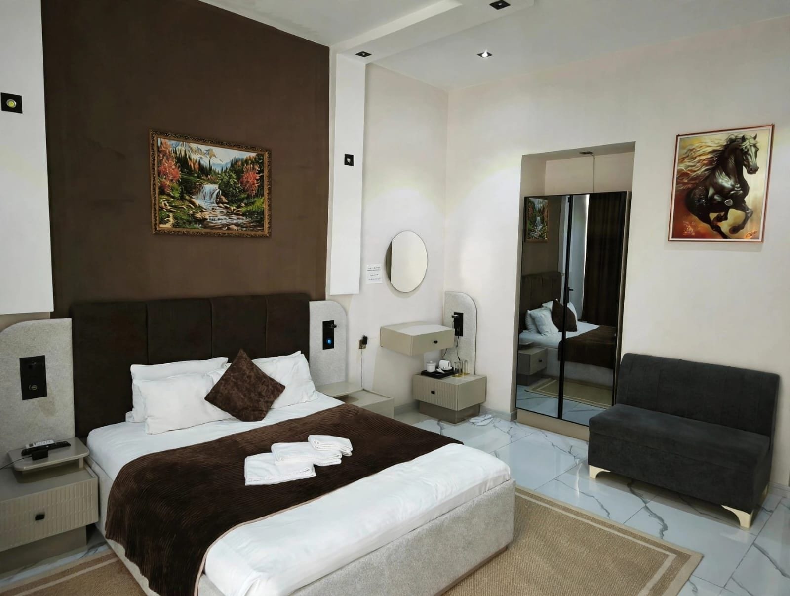 Habitación de hotel con una cama grande, mesita de noche, espejo, zona de estar y obras de arte en las paredes.