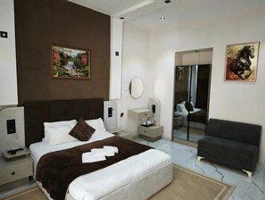 Quarto de hotel com uma cama grande, mesa de cabeceira, espelho, área de estar e obras de arte nas paredes.
